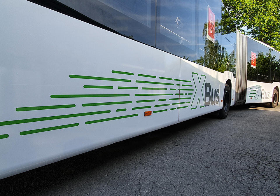 Weißer Gelenkbus von der Seite. Auf dem Bus sind grüne Streifen zu sehen, die vor und hinter dem Gelenk ein großes X bilden. Dahinter steht jeweils das Wort „Bus“ in Grau.