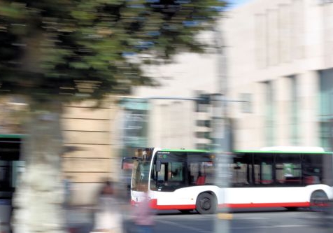 Ein Bus fährt durch die Stadt. Bäume, weitere Busse und Personen im Hintergrund sind durch die Bewegung verschwommen.