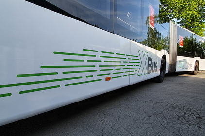 Weißer Gelenkbus von der Seite. Auf dem Bus sind grüne Streifen zu sehen, die vor und hinter dem Gelenk ein großes X bilden. Dahinter steht jeweils das Wort „Bus“ in Grau.