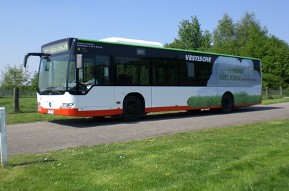 Ein Bus der Vestischen fährt bei sonnigem Wetter auf einer Straße durch eine grüne Wiesenlandschaft.