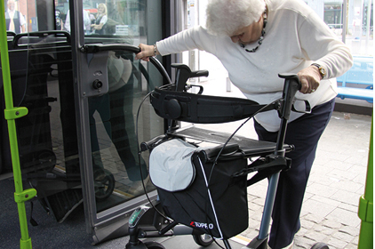 Eine Frau steigt mit ihrem Rollator rückwärts aus dem Bus. Dabei hält sie sich am Handlauf fest.